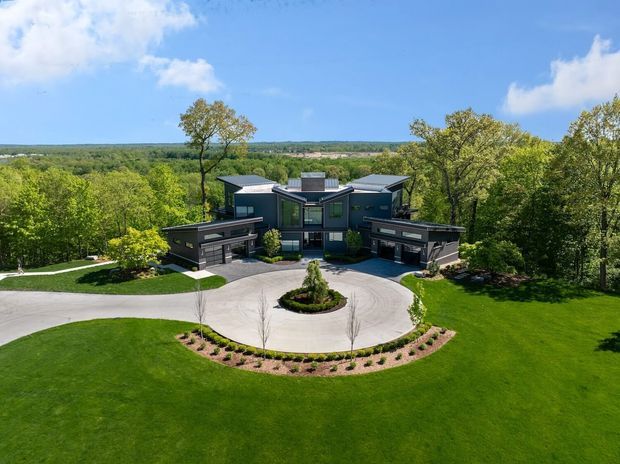 Aerial exterior view of a large contemporary dark-clad residence set within expansive landscaped grounds and mature woodland. The home has a strongly symmetrical composition with projecting wings, multiple low-pitched roof planes, broad glazing, and a circular driveway with a central planted island. Manicured lawns and curved garden beds frame the approach, with a wooded rural landscape extending beyond.