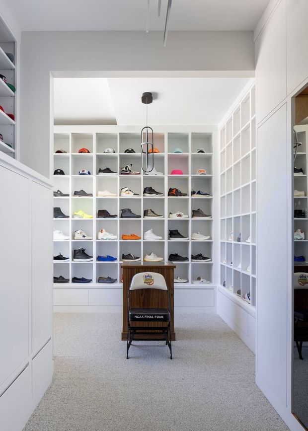 Bright, highly organised walk-in wardrobe or dressing room with extensive white built-in open shelving displaying a large collection of shoes and caps. The room has white cabinetry, glass-fronted display storage along the right wall, carpet flooring, a central timber sports chair, and a minimalist pendant light. A mirror or reflective panel on the right reinforces the sense of built-in storage and enlarges the space visually.