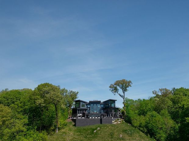 Exterior view of a large contemporary multi-level house set on a wooded, elevated or sloping site. The dark facade features extensive glazing, projecting upper volumes, terraces or balconies, and a substantial dark retaining or foundation wall. Dense green trees surround the property beneath a clear blue sky.