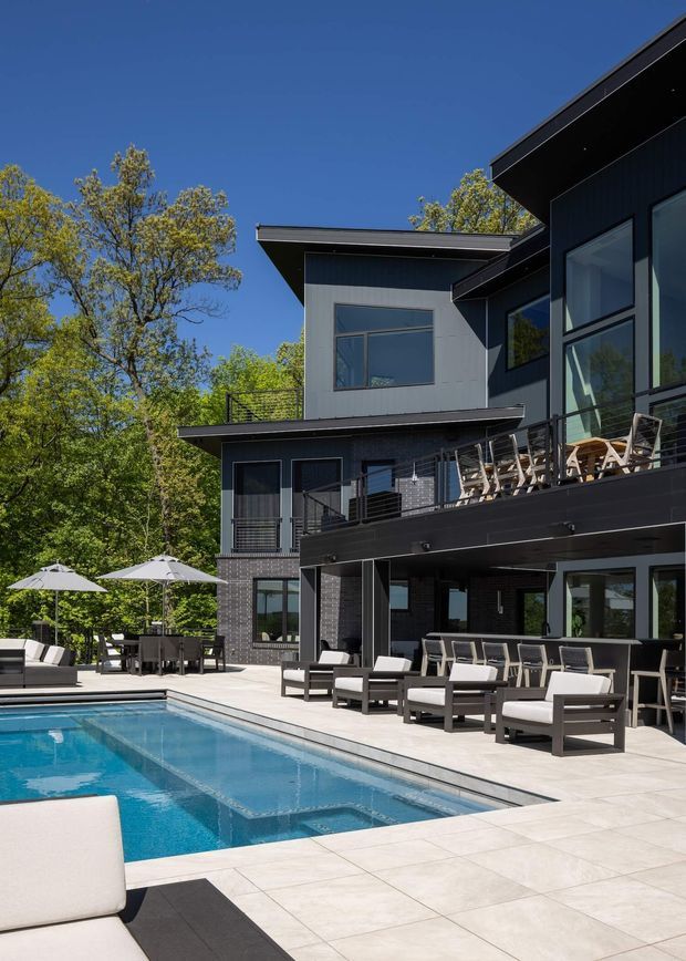A contemporary multi-level dark-clad residence viewed from its pool terrace. The home features strong horizontal roof overhangs, extensive glazing, balconies with slim metal balustrades, and a covered outdoor entertaining area. A rectangular swimming pool is surrounded by pale tiled paving, lounge furniture, dining settings, and umbrellas, with mature trees beyond the property.