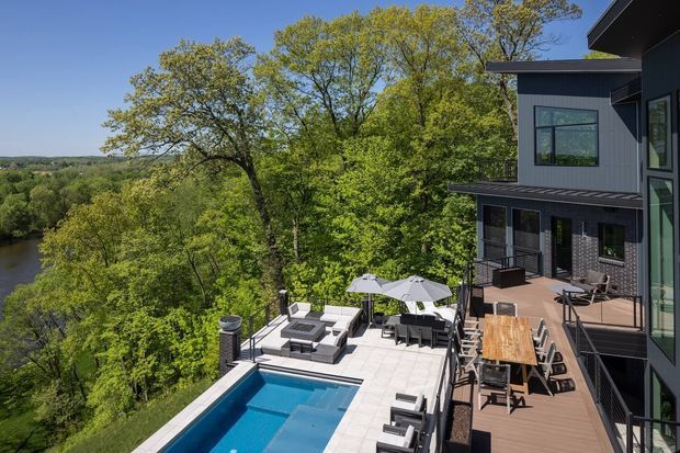 Elevated exterior view of a contemporary multi-level house overlooking a wooded river or valley landscape. The image shows dark vertical cladding, extensive glazing, projecting roof forms, a timber deck with outdoor dining, a paved entertaining terrace, and a swimming pool.