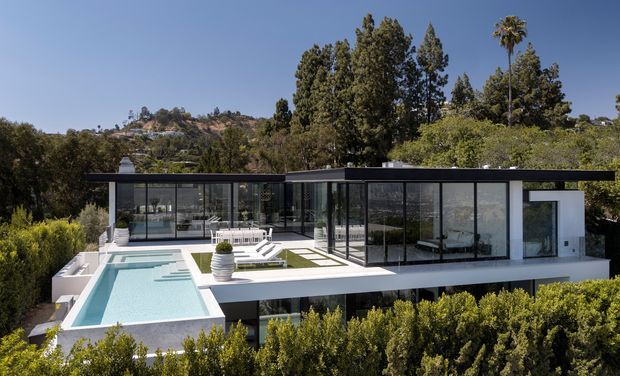 Exterior view of a modern, flat-roofed multi-level residence set into a leafy hillside. The house features extensive floor-to-ceiling glazing, crisp white rendered walls, dark slender roof and window frames, broad terraces, and a long rectangular swimming pool with integrated outdoor lounging areas.