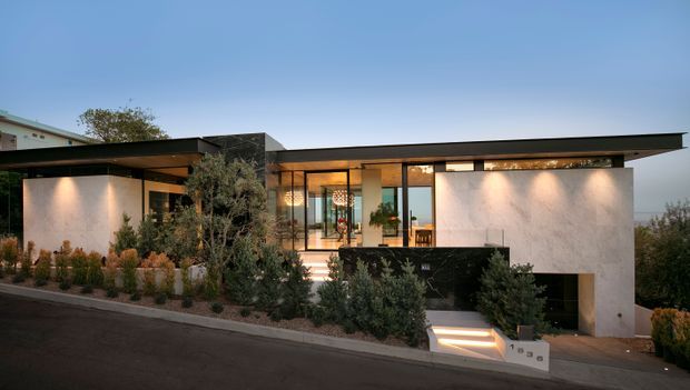 A dusk exterior view of a low-slung contemporary residence on a sloping site. The house features broad flat roof planes with deep overhangs, white rendered walls, dark stone or masonry feature volumes, extensive floor-to-ceiling glazing, and a landscaped frontage with a stepped entry descending to a lower-level opening.