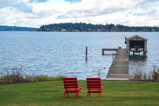 A waterfront residential landscape viewed from a neatly maintained lawn, with two red outdoor chairs facing a calm lake. A timber dock extends into the water toward a small covered boat house, with wooded shoreline and distant homes across the lake.