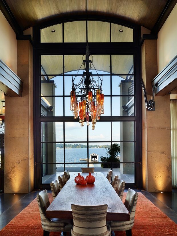 A dramatic double-height dining room with a long timber dining table and upholstered chairs arranged before expansive black-framed floor-to-ceiling glazing. A large sculptural cluster chandelier hangs centrally beneath a curved timber-lined ceiling. Warm stone or plaster walls, dark timber framing, and a deep red rug create a refined, richly textured interior. Through the glazing, a waterfront, dock, and opposite shoreline are visible.