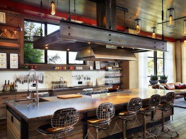 A spacious, industrial-inspired kitchen with a large central island and seating for five. The kitchen features dark timber cabinetry and wall panelling, a warm wood ceiling, exposed dark structural beams, a tiled splashback, stainless-steel appliances and sink fittings, and a substantial suspended metal range hood. Pendant lights hang above the island, while large windows provide abundant natural light. A living area is visible beyond the kitchen.