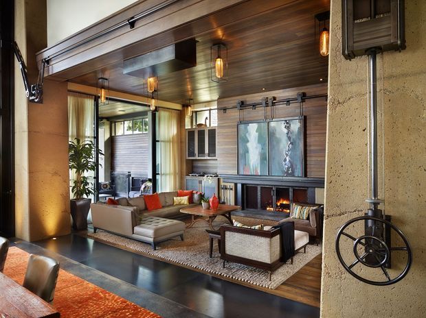 Industrial-style living room with a warm timber-lined ceiling and feature wall, exposed concrete columns, black metal detailing, and a large glazed garage-style opening. The room contains a sectional sofa, lounge chairs, a low timber coffee table, built-in cabinetry, artwork, and a contemporary fireplace. Warm pendant lights and orange accents create a cozy, refined atmosphere, while the open glazing connects to a planted outdoor area.