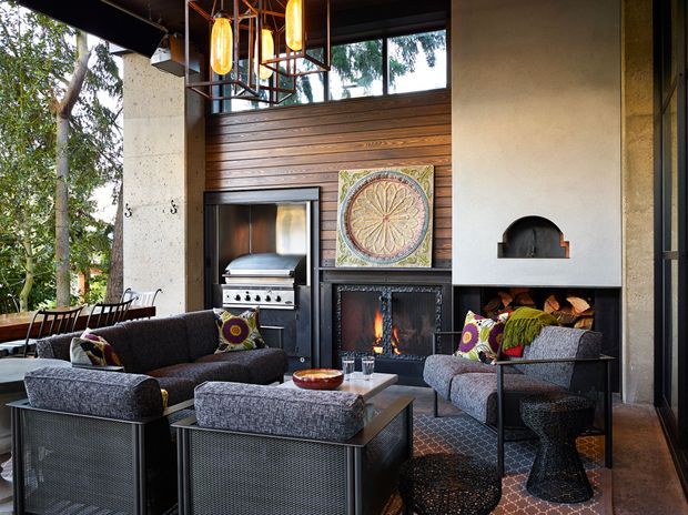 A sophisticated indoor-outdoor living space with a high ceiling, exposed concrete columns, extensive glazing, and a dramatic fireplace wall. The room includes two dark upholstered sofas, metal-framed occasional tables, a built-in gas grill, timber wall panelling, patterned tiled flooring, and sculptural pendant lights. A decorative circular artwork is mounted above the fireplace, which has integrated firewood storage and a small arched niche.