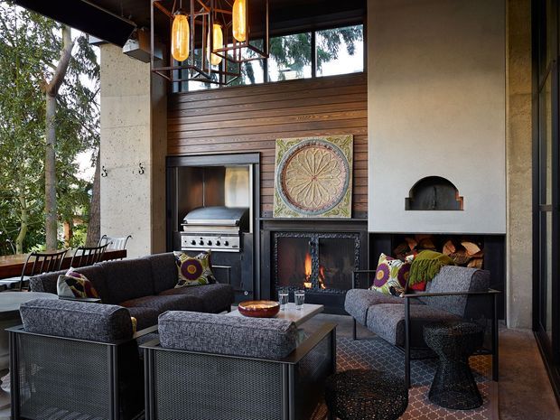 A sophisticated indoor-outdoor living and entertaining space with a central fireplace, built-in barbecue, timber wall panelling, exposed concrete columns, and large glazed openings overlooking greenery. Dark upholstered sofas and armchairs surround a low coffee table, while warm pendant lights and patterned cushions add an inviting, eclectic character.