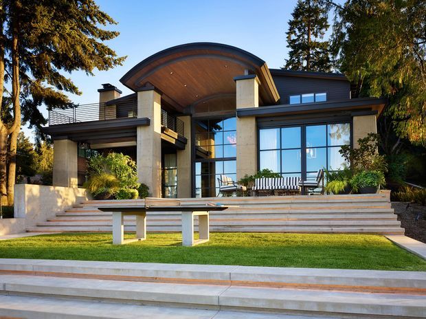 Exterior view of a sophisticated contemporary multi-level home set among mature trees. The facade combines dark horizontal cladding, pale stone or concrete piers, extensive floor-to-ceiling glazing, and a prominent curved timber-lined roof canopy. Broad tiered steps lead to a raised terrace and manicured lawn, with outdoor seating and integrated landscaping.