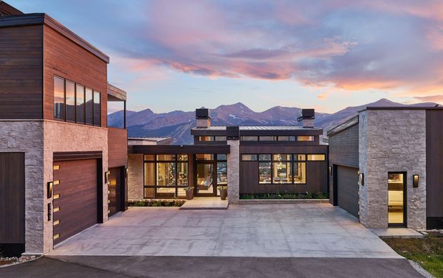 Exterior view of a contemporary multi-building residence arranged around a broad paved driveway or courtyard. The main facade combines warm horizontal timber cladding, grey stone masonry, dark metal-framed glazing, flat roof forms, and prominent garage and entry doors. Mountain ridges are visible beyond the property under a colourful sunset sky.