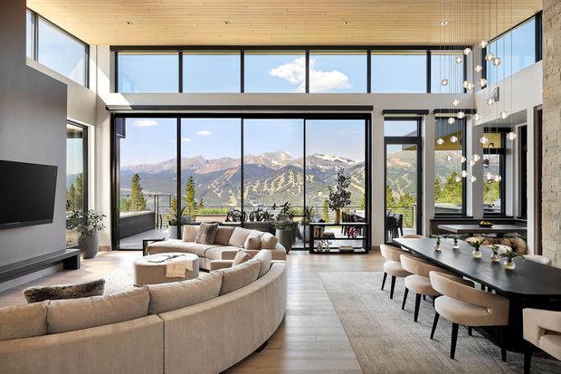 A spacious, light-filled open-plan living and dining area with expansive floor-to-ceiling glazing overlooking a dramatic mountainous landscape. The room features a large curved sectional sofa, minimalist fireplace and media wall, a long dark dining table with upholstered chairs, extensive built-in glazing, a warm timber-lined ceiling, pale timber flooring, and a sculptural cluster pendant above the dining area. A stone-clad wall adds natural texture.