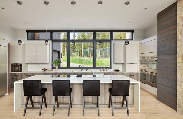 A spacious contemporary kitchen with a large central island and four dark upholstered bar stools. The room features white flat-panel cabinetry, integrated stainless-steel appliances, a timber-look island base, pale timber flooring, and extensive black-framed glazing overlooking a wooded landscape. A dark horizontal timber feature wall and adjacent stone-clad column add contrast and texture. Multiple pendant lights hang above the island.