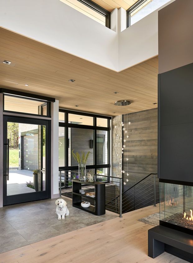 A bright, contemporary open-plan entry and living area with a dramatic double-height void, clerestory glazing, black-framed windows and doors, and a timber-lined ceiling. Large-format grey floor tiles transition to pale timber flooring near a freestanding contemporary fireplace. A black metal stair balustrade overlooks a lower level, while a console display unit and decorative pendant lights sit near the glazed entry. The space is airy and refined, with strong indoor-outdoor connection through extensive glazing.