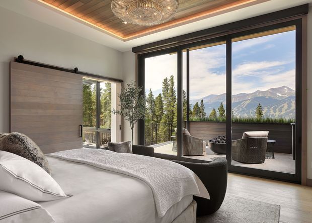 A luxurious bedroom with a large bed in the foreground, a warm timber-lined recessed ceiling with integrated lighting, and a sculptural chandelier. Full-height dark-framed glazing opens to a balcony or terrace furnished with lounge chairs and a small table, offering views of a wooded mountain landscape. The room uses a soft neutral palette with timber and dark metal accents.