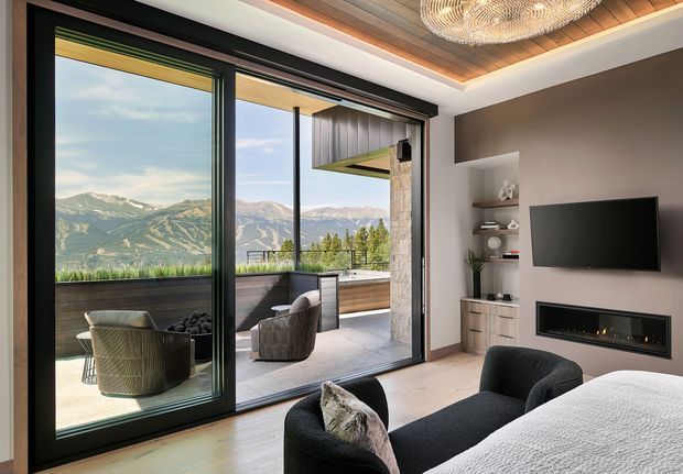 A contemporary bedroom with a large bed in the foreground, a dark upholstered lounge chair, wall-mounted television, built-in shelving, and a linear gas fireplace. Full-height black-framed sliding glass doors open to a furnished balcony or terrace with expansive mountain views. The room features light timber flooring, a warm timber ceiling detail with recessed lighting, and a sculptural woven pendant light.