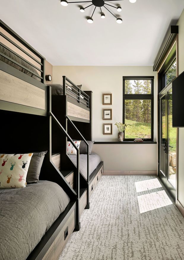 A bright, compact bunk bedroom with two built-in bunk beds along the walls. The bunks combine dark charcoal framing with pale timber panels and integrated storage drawers. A large black-framed window and glazed door bring in abundant daylight and views of a wooded landscape. The room has a pale neutral wall, a textured light-grey carpet, framed artwork, grey bedding, and a modern black multi-arm ceiling light.