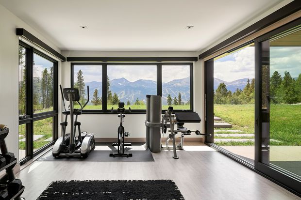 A bright, modern home gym enclosed by expansive black-framed glazing, with cardio and strength-training equipment arranged on a pale timber-look floor. The room opens visually and physically toward a grassy outdoor area and mountain landscape, creating an airy indoor-outdoor connection. A dark structural beam and recessed ceiling lights add architectural definition, while a black rug provides contrast.