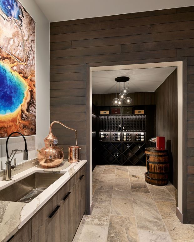 A refined home bar and wine storage area viewed through a wide timber-lined opening from an adjacent kitchenette or butler's pantry. The foreground includes a deep stainless-steel sink, pale stone benchtop, dark cabinetry, copper-toned decorative vessels, and a large colourful abstract artwork. Beyond, a dark built-in wine cellar displays bottles and glassware beneath clustered pendant lights, with a wooden barrel used as a side table. Large-format stone-look flooring continues through both spaces.