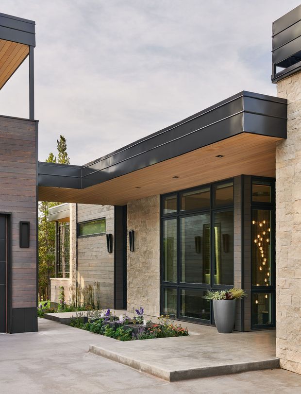 Contemporary residential entrance with a flat-roofed projecting canopy, dark metal fascia, warm timber soffit, pale stone-clad walls, and dark-framed glazing. A recessed glass front door opens onto a broad concrete entry terrace, with linear garden beds and architectural planting along the facade.