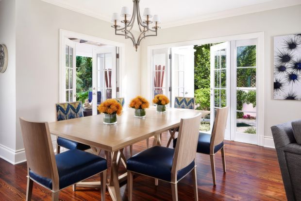 Bright, elegant dining room with a large rectangular timber dining table and upholstered dining chairs, opening through French doors to a leafy outdoor area. The room has white walls and trim, polished dark timber flooring, a decorative chandelier, patterned accent cushions, and botanical artwork.