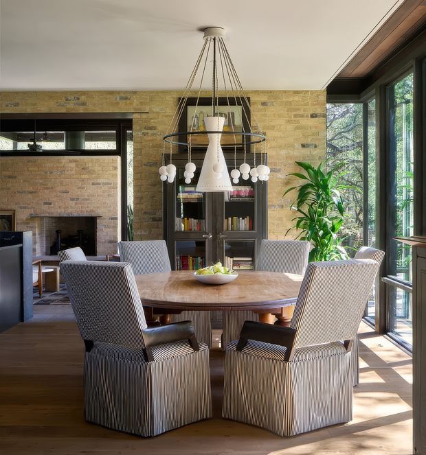 Warm, light-filled dining room with a round timber dining table and six upholstered slipcovered chairs. The room features exposed tan brick walls, dark timber built-in shelving, a large sculptural pendant light, timber flooring, and extensive floor-to-ceiling glazing overlooking greenery. A fireplace and adjacent living area are visible at left.
