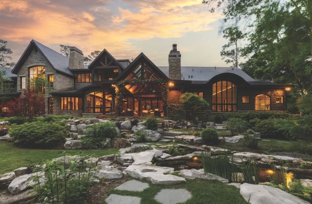 A large multi-wing residential home viewed from a landscaped garden at dusk. The house features steep gabled roofs, dark timber framing, grey stone or shingle-clad walls, extensive warm-lit glazing, a prominent stone chimney, and a curved-roof wing. The foreground contains a naturalistic rock garden, pond, stepping stones, shrubs, and mature trees.