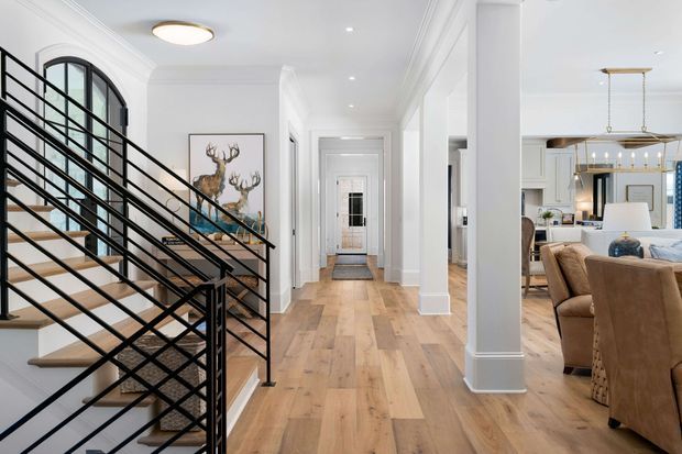Bright, refined residential interior showing a wide hallway and entry circulation space with a timber staircase on the left. The staircase has black metal balustrades and light timber treads, while the hall features white walls, substantial decorative architraves and skirting, light timber flooring, recessed lighting, and large square columns. An open-plan dining and living area with upholstered dining chairs and pendant lighting is visible to the right.