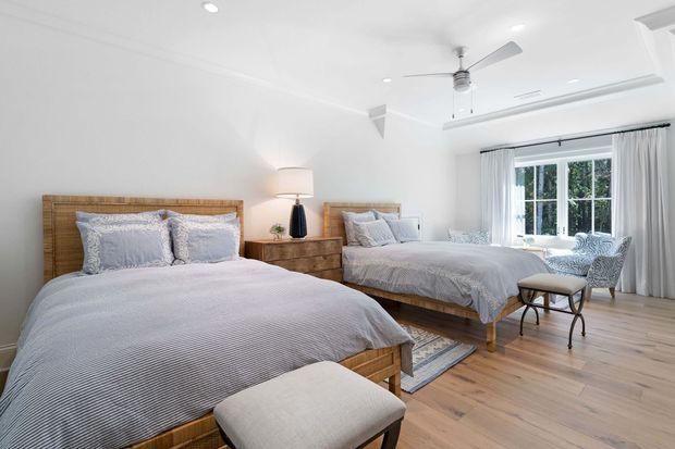 Bright, spacious bedroom with two beds, woven timber headboards, pale textured bedding, timber bedside furniture, a table lamp, upholstered bench, patterned armchair, and light timber flooring. White walls and ceiling, recessed lighting, a ceiling fan, and full-height curtains surround a large window overlooking greenery. The room has a calm, airy coastal-traditional feel.