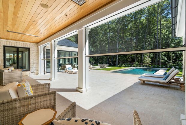 A bright, enclosed outdoor living space with large retractable glazed openings overlooking a swimming pool and landscaped grounds. The room features woven rattan seating, pale stone paving, a white-painted masonry wall, and a warm timber-lined ceiling with recessed lighting. The open glazing connects the lounge directly to the pool terrace and garden.