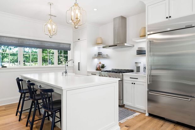 Bright, refined white kitchen with a large waterfall-edge island, integrated seating for four, white shaker-style cabinetry, stainless-steel range hood and refrigerator, open shelving, and warm timber flooring. A wide bank of windows brings in abundant natural light, while two decorative brass-and-glass pendant lights add a traditional accent.