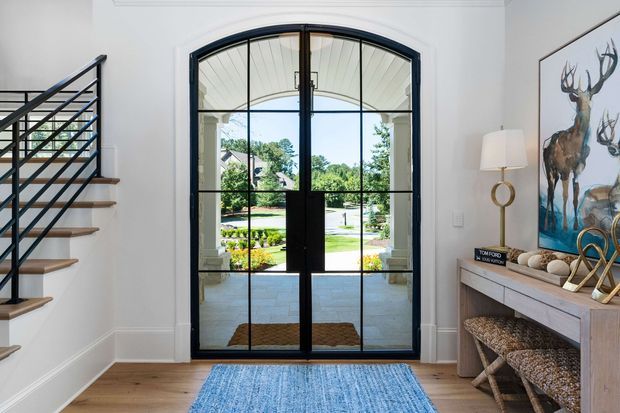 Bright, refined entry foyer viewed from inside, featuring a tall arched black steel-framed glazed front door with divided panes, white walls and trim, a warm timber floor, and a staircase with black horizontal railing and timber treads. A slim console table with stools, lamp, artwork, and decorative objects occupies the right side. The doorway opens to a landscaped exterior and covered porch.
