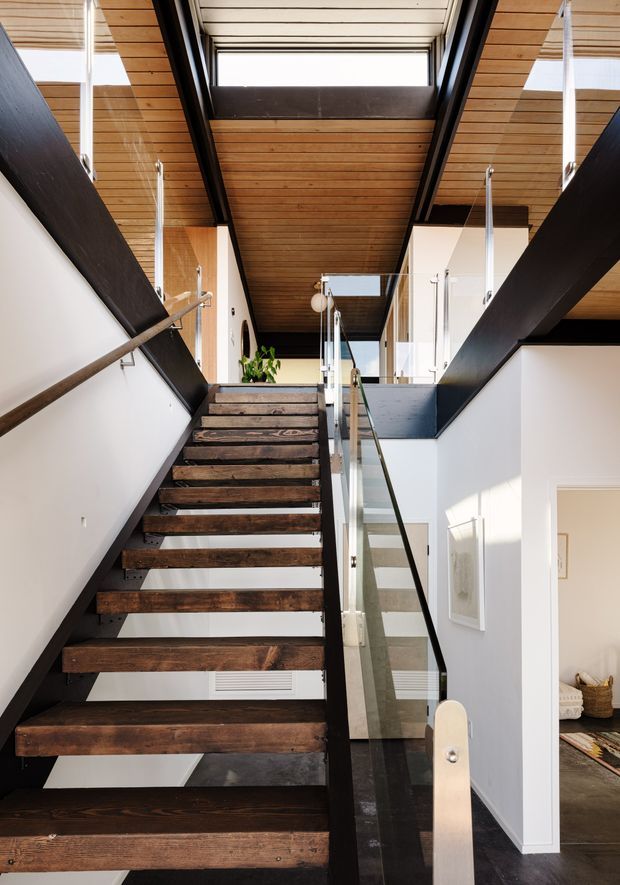 Interior view looking up a modern staircase with open dark timber treads, black structural stringers, and clear glass balustrades with metal handrails. White walls contrast with a warm timber-lined ceiling and large clerestory/skylight glazing, creating a bright, vertically connected stair hall. An upper-level landing and part of an adjacent room are visible.