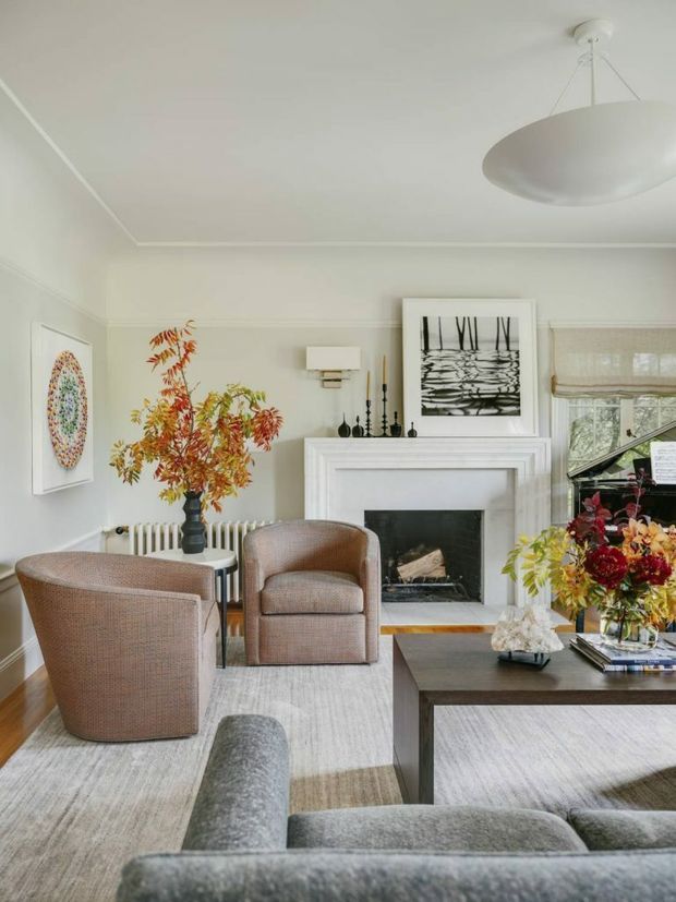 Bright, refined living room with a white-painted fireplace mantel, pale walls with decorative moulding, a large sculptural pendant light, framed artwork, woven upholstered armchairs, a grey sofa, and a dark rectangular coffee table. Autumnal floral arrangements and neutral furnishings add warmth to the otherwise light, classic interior.