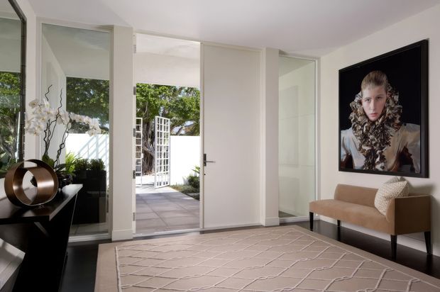 Bright contemporary entry hall with white walls, full-height glazed doors and sidelights opening to a landscaped courtyard. A slim black console with sculptural decor sits to the left, while a tan upholstered bench and large framed artwork create a refined seating vignette on the right. The room has dark timber flooring, a pale patterned area rug, and abundant natural light.
