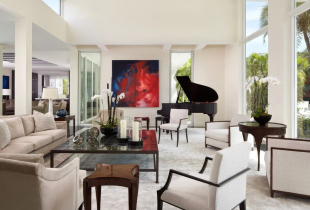 A bright, spacious contemporary living room with very high ceilings and expansive floor-to-ceiling windows. The room features a large cream sectional sofa, white upholstered lounge chairs, a dark glass-topped coffee table, a black grand piano, a sculptural red-and-blue artwork, indoor plants, and a pale patterned area rug. An adjacent dining or sitting area is visible through the open connection on the left, while lush palms are visible outside.