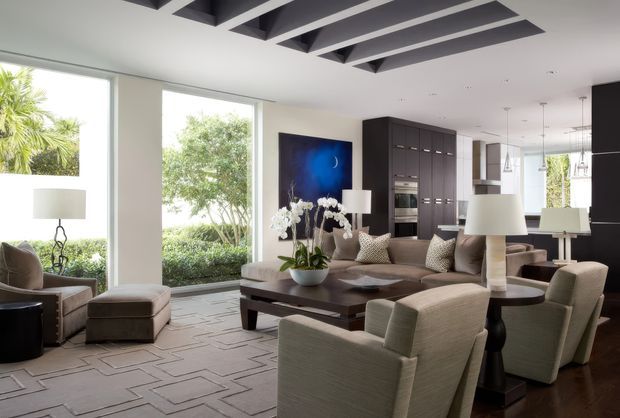 Sophisticated open-plan living and dining area with large floor-to-ceiling windows, a neutral upholstered seating arrangement, dark timber coffee table, sculptural lamps, dark built-in cabinetry, and a kitchen visible in the background. The ceiling features bold dark exposed beams or recessed geometric detailing, while an abstract blue artwork and orchid arrangement provide accents.
