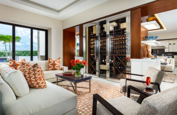 Bright, refined living room centered on a large built-in glass-front wine storage wall, with white upholstered seating, dark wood-framed armchairs, a glass coffee table, patterned rug, and orange patterned cushions. The room opens visually toward a white kitchen and dining area, while large windows provide abundant natural light and views of greenery and water beyond. A recessed tray ceiling and extensive dark timber cabinetry add architectural definition.