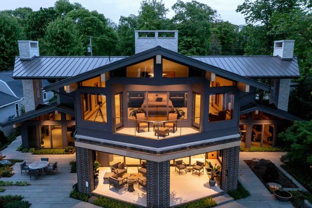 A large multi-level contemporary residence viewed from the front at dusk, set within mature trees. The house features a dramatic central gabled roof with broad metal roof planes, multiple masonry-clad chimneys, extensive warm-lit glazing, dark structural framing, and a projecting two-storey central pavilion. Open upper and lower outdoor living areas with seating are visible, connected by paved paths and landscaped planting.