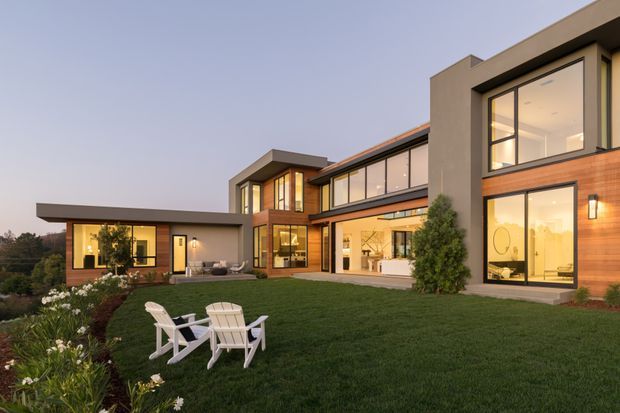 A contemporary multi-wing residential house viewed from a landscaped lawn at dusk. The home has strong geometric volumes, extensive floor-to-ceiling glazing, warm timber wall cladding, smooth beige-grey rendered walls, dark roof planes, and illuminated interiors. White outdoor lounge chairs sit on the lawn, with garden beds and shrubs around the property.