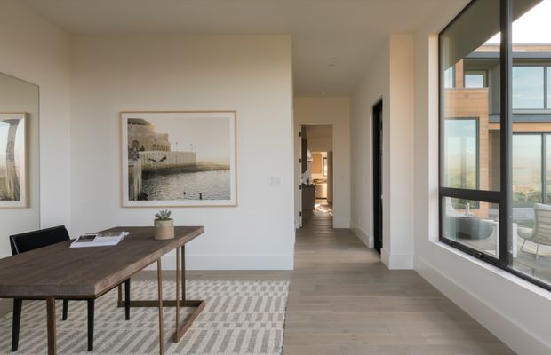 Bright, understated contemporary dining area in an open-plan interior. A timber dining table with black chairs sits on a patterned rug beside large black-framed floor-to-ceiling windows, with pale walls, light timber flooring, framed artwork and a hallway leading toward the kitchen or living spaces. An adjacent modern building and landscape are visible through the glazing.