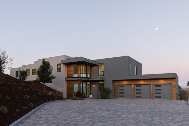 Contemporary multi-level residential house photographed from the front at dusk. The facade combines dark charcoal rendered walls with white rendered volumes, extensive floor-to-ceiling glazing, warm timber-toned entry detailing, and an attached three-car garage. A broad paved driveway and landscaped sloping garden occupy the foreground.