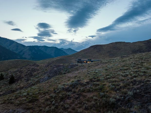 A small, low-profile building is nestled into a rugged alpine hillside, surrounded by open tussock vegetation. Layered mountain ranges and a dramatic cloudy sky form the background. The structure appears to have a dark, gently sloping or flat roof with broad glazing, creating a strong connection to the remote landscape.