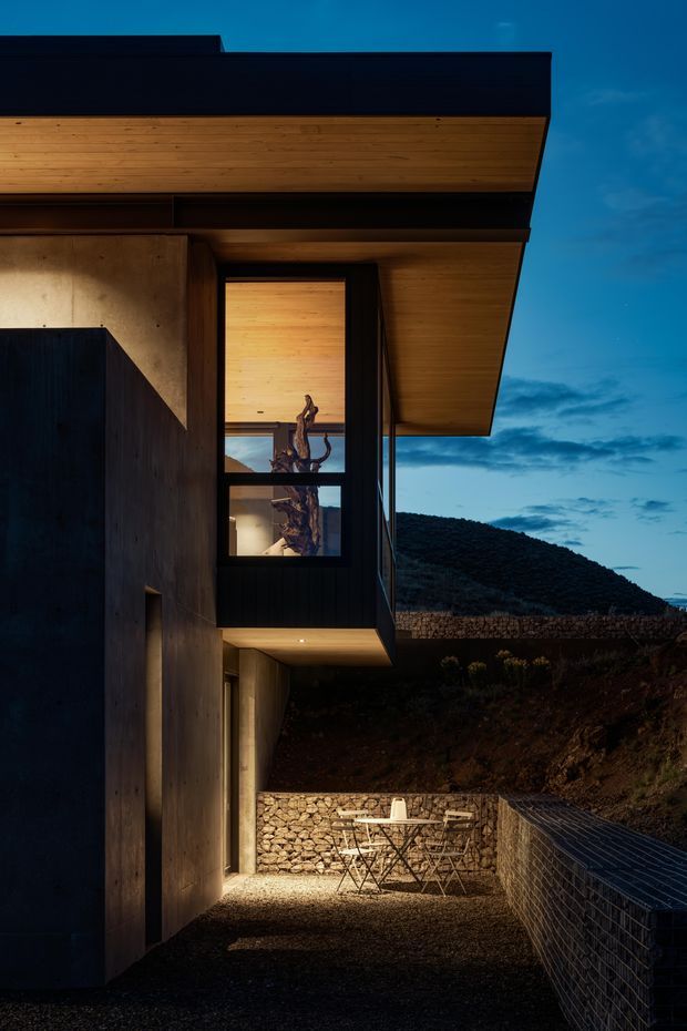 A dramatic dusk exterior view of a modern hillside dwelling, focused on a cantilevered upper volume with a deep timber-lined soffit and dark roof edge. The facade combines raw concrete-toned walls, dark vertical cladding, and large black-framed glazing. A warmly lit interior and sculptural branch-like feature are visible through the window. Below, a gravel terrace with a small outdoor dining set is enclosed by gabion stone retaining walls, with a rugged wooded hillside beyond.
