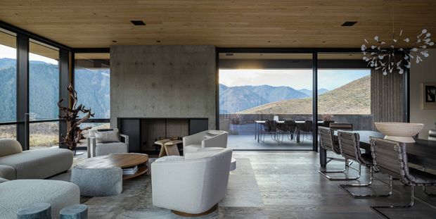 A sophisticated open-plan living and dining area with expansive floor-to-ceiling glazing overlooking rugged mountainous terrain. The room features a sculptural concrete fireplace wall, warm timber-lined ceiling, dark-framed glazing, pale upholstered lounge seating, a large dining table with contemporary cantilever chairs, polished grey flooring, and a statement branching pendant light. An outdoor terrace is visible beyond the glazing.