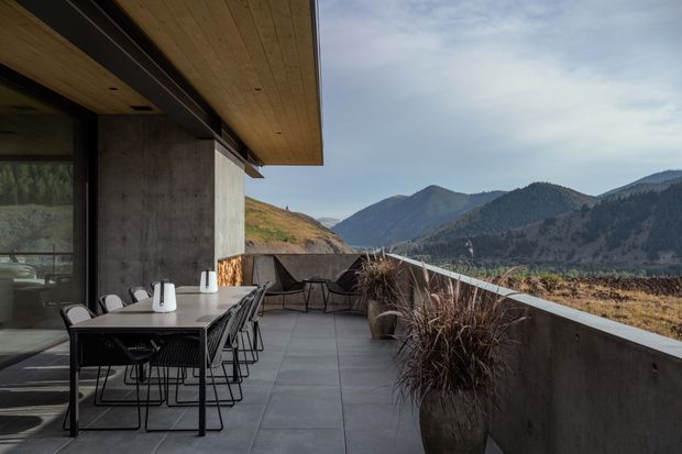 A contemporary mountain-view terrace adjoining a modern concrete building. The spacious outdoor dining area has a dark rectangular table with black wire-frame chairs, large grey tiled paving, a concrete privacy wall and balustrade, and timber soffit overhead. Dried ornamental grasses in planters line the edge, with expansive forested mountains visible beyond.
