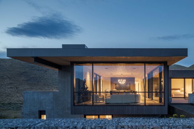 Twilight exterior view of a contemporary single-storey residence set into a sloping, rocky site. The house has a strong flat roof, dark horizontal cladding, expansive floor-to-ceiling glazing, and a projecting timber-lined soffit. Warm interior lighting reveals an open-plan living area through the glass, while concrete walls, stone landscaping, and a partially visible adjoining wing reinforce the minimalist architectural composition.
