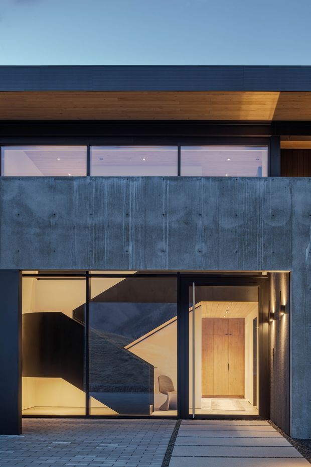 Exterior architectural photograph of a contemporary, multi-level residence at dusk. The facade combines dark horizontal cladding, warm timber soffits and wall lining, exposed concrete, and extensive dark-framed glazing. A recessed entry is visible at ground level, opening into a warmly lit interior with timber joinery, while a paved approach leads toward the doorway. Strong geometric volumes, deep overhangs, and contrast between cool concrete and warm wood define the composition.