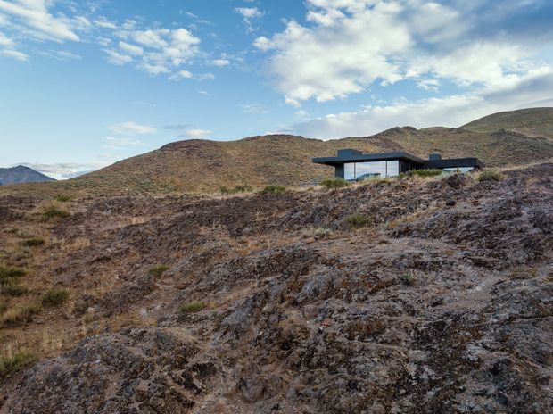 A low-profile modern house with extensive glazing and a dark flat roof sits partially embedded in a rugged, treeless mountainous landscape. The wide view emphasizes the rocky terrain, surrounding hills, and expansive blue sky.