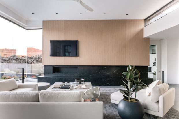 Bright, refined contemporary living room with a large cream sectional and armchairs arranged around a low coffee table. A full-height light timber feature wall incorporates a wall-mounted television, while a dark veined stone fireplace surround or feature band anchors the room. Floor-to-ceiling glazing opens toward an outdoor area, and recessed ceiling lights and a ceiling fan contribute to the clean modern fit-out.